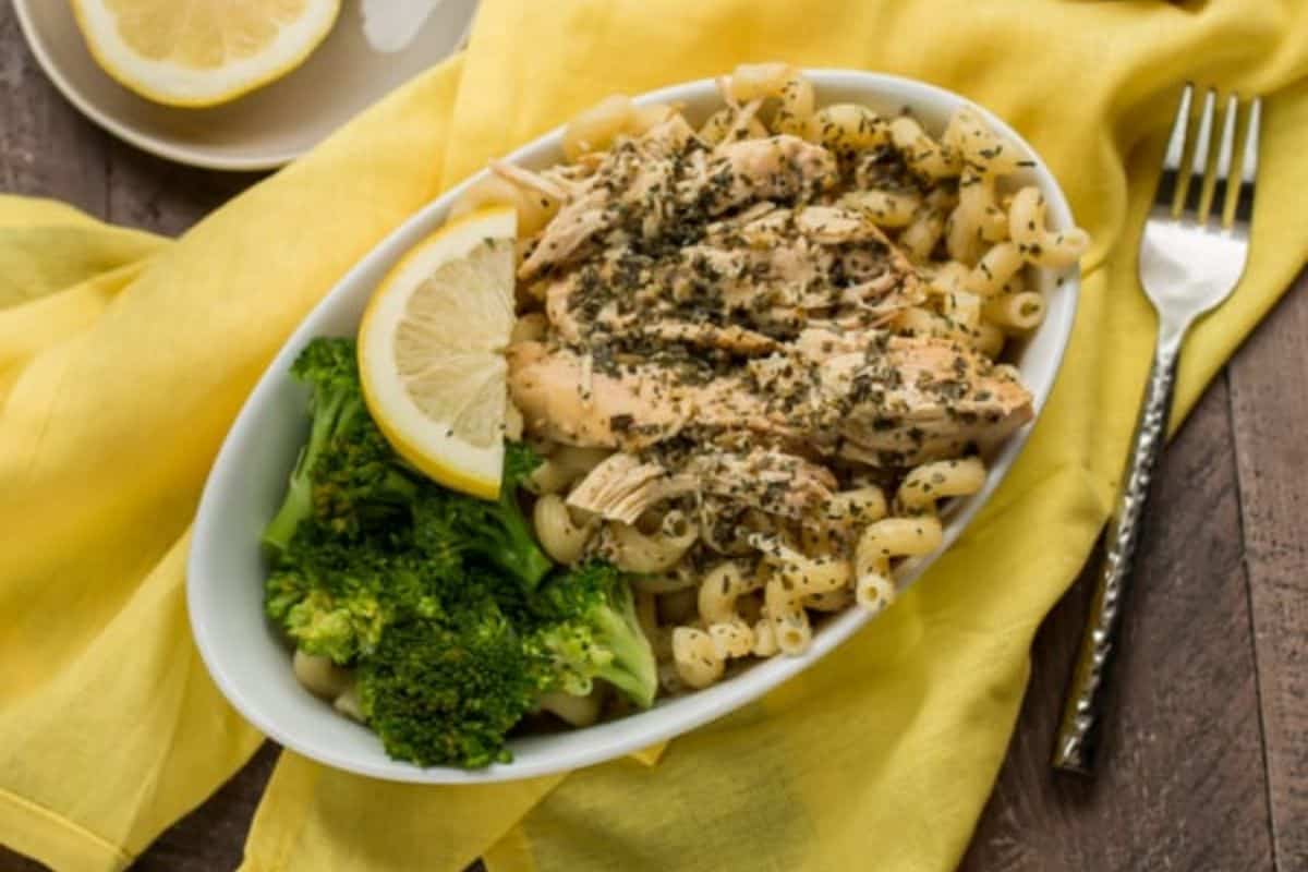 A white bowl with cooked pasta, seasoned chicken, steamed broccoli, and a lemon slice, placed on a yellow cloth next to a fork—perfect for easy new mom freezer meals.