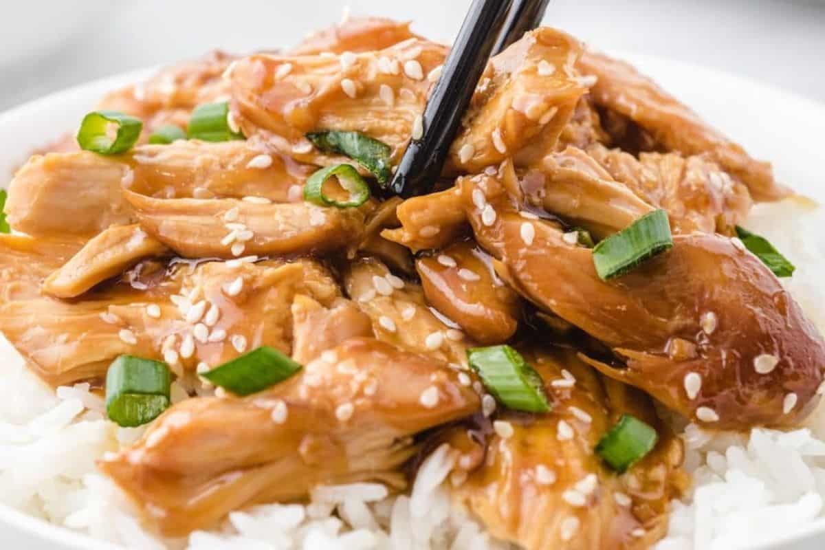 A bowl of white rice topped with shredded chicken in brown sauce, garnished with sesame seeds and chopped green onions—one of the perfect new mom freezer meals—served with chopsticks picking up a piece.