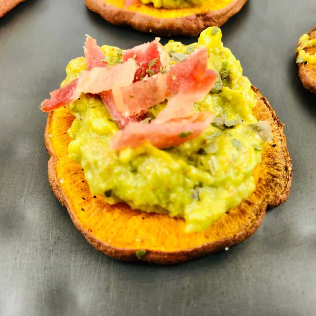 A delicious Sweet Potato Appetizer featuring a slice of roasted sweet potato topped with creamy mashed avocado and thin strips of bacon, elegantly served on a dark surface.