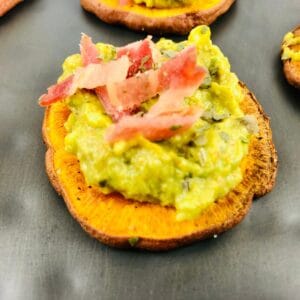 A delicious Sweet Potato Appetizer featuring a slice of roasted sweet potato topped with creamy mashed avocado and thin strips of bacon, elegantly served on a dark surface.
