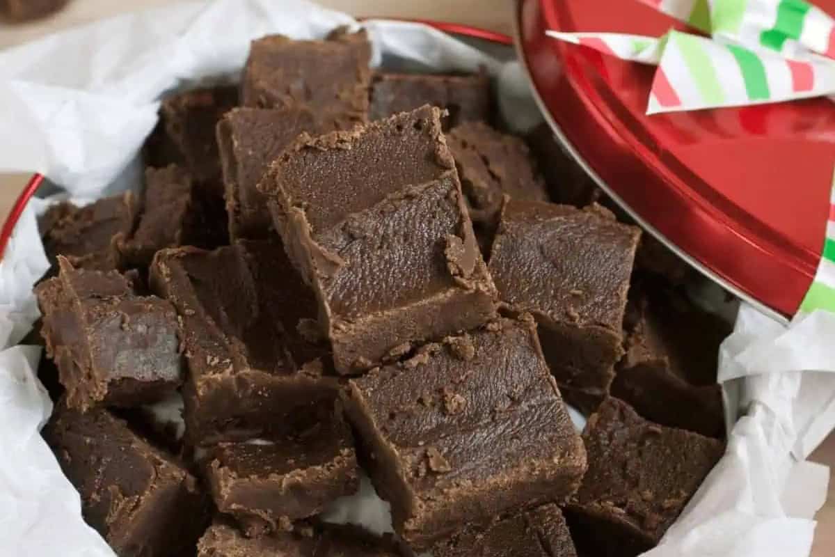 A tin filled with neatly stacked squares of 2 ingredient fudge on white parchment paper, with the tin's red lid partially visible.