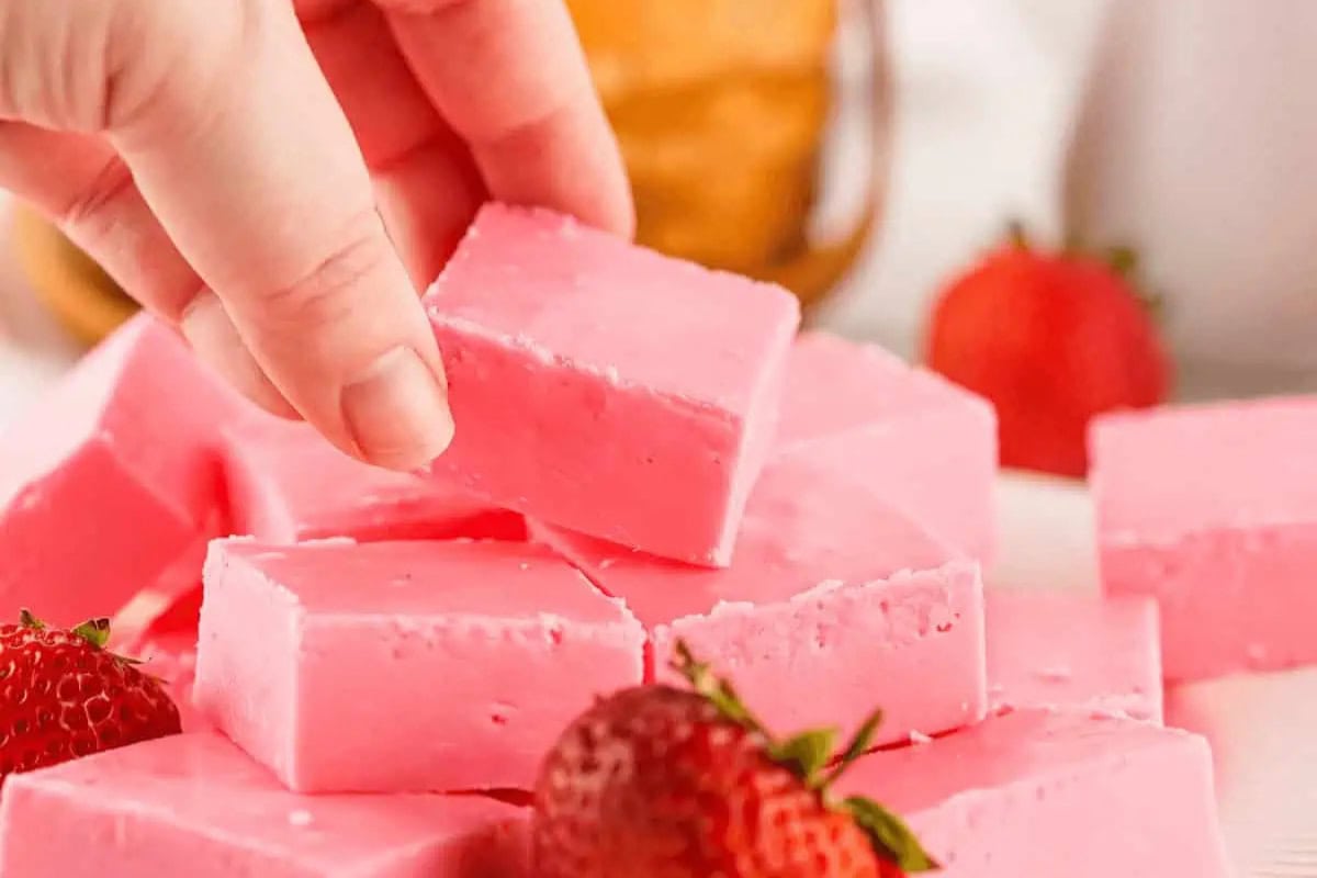 A hand picks up a piece of pink strawberry fudge from a pile, with fresh strawberries placed nearby.