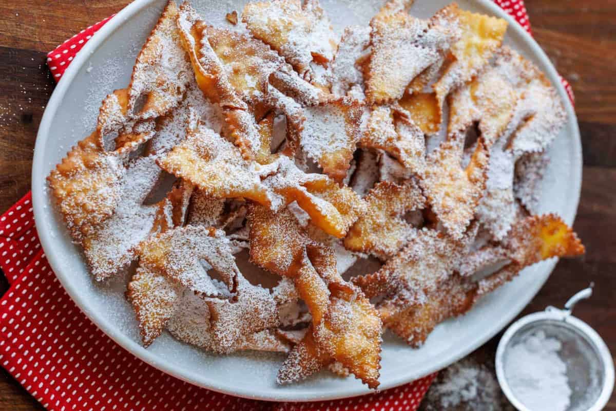 A white plate of fried angel wing pastries, a classic Russian Christmas food, dusted with powdered sugar sits on a red polka dot napkin, with a small strainer of powdered sugar nearby.