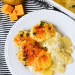 Plate of the side dish, garnished with herbs, next to cubes of cheddar and a whole potato, on a striped cloth.