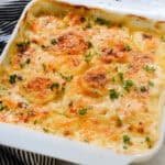 A baked dish of Au Gratin Potatoes with Gruyere Cheese in a white square dish, topped golden brown and garnished with chopped parsley, rests on a black and white striped cloth on a marble surface.