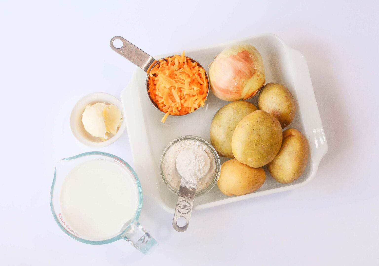 A tray with potatoes, onion, cheese, a measuring cup with milk, a measuring spoon with flour, and a small bowl of butter.