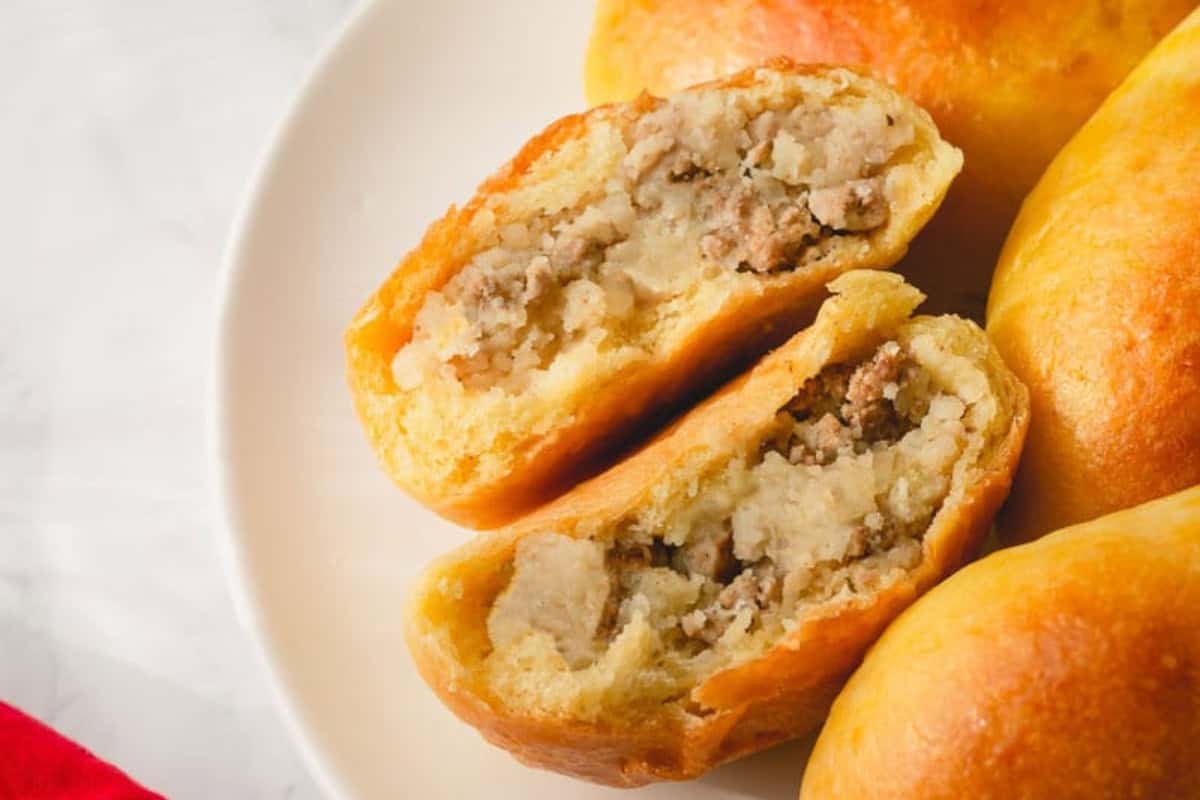A close-up of a plate with baked piroshki meat pies, one cut open into two halves to show a filling of ground meat and potatoes/