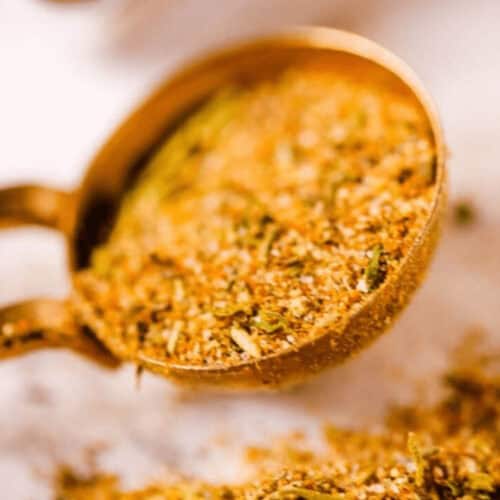 A close-up of a measuring cup filled with a blend of dried herbs and spices, some spilling out onto the surface below, perfect for creating a savory chicken rub for smoking.