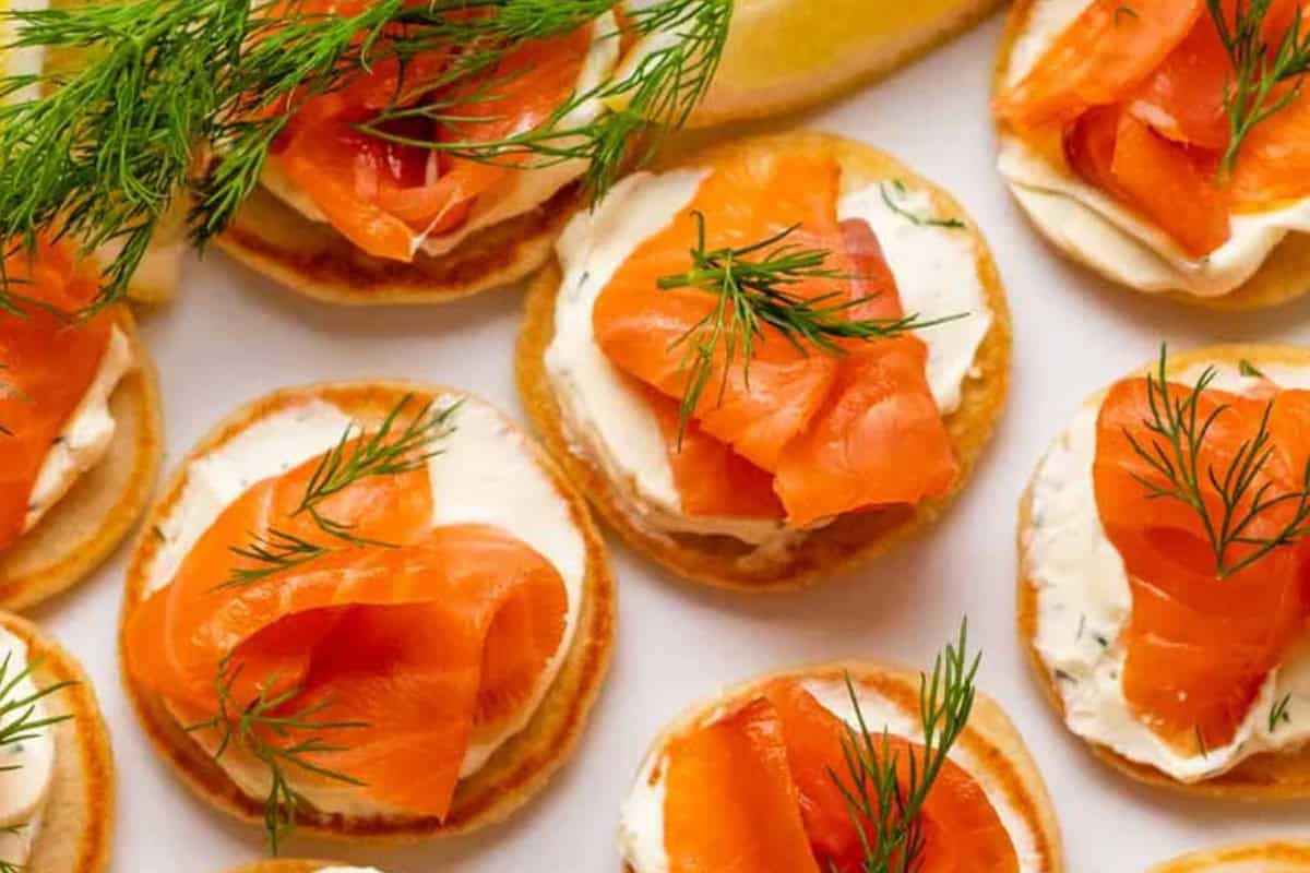 Small blinis, a classic Russian food, are topped with white crème fraîche, smoked salmon slices, and fresh dill sprigs and arranged on a white surface.