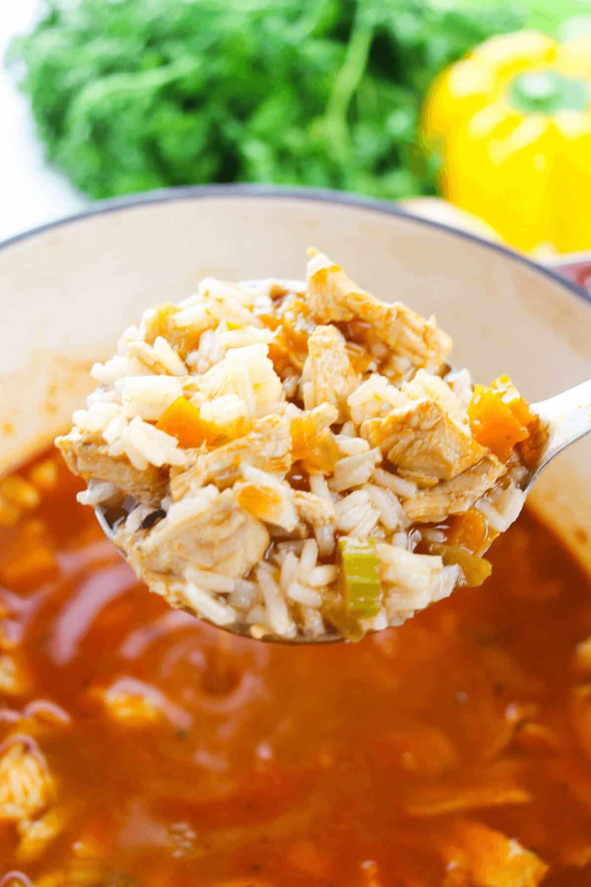 A ladle holding a portion over a pot reveals tender chicken, rice, and vibrant vegetables. Fresh herbs and a yellow bell pepper blur softly in the background, enhancing this flavorful Cajun delight with aromatic charm.