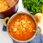 A bowl of savory tomato rice soup with a hint of Cajun flair sits invitingly in front of a pot. Fresh parsley, onion, and a celery stalk enhance the flavors, evoking the essence of Cajun chicken and rice dishes.