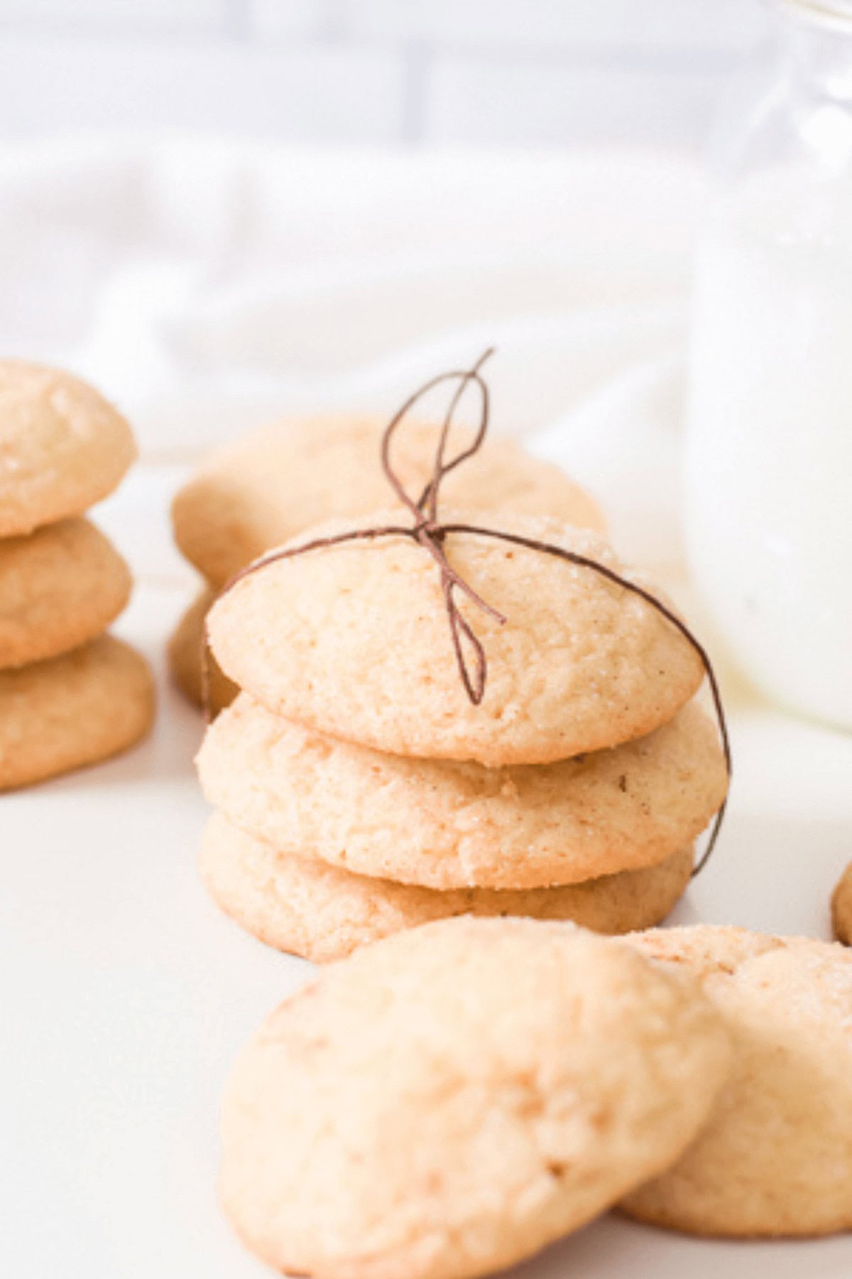 A stack of round snickerdoodle cookies made with cake mix is tied with twine, surrounded by more cookies. A glass bottle of milk sits invitingly in the background.