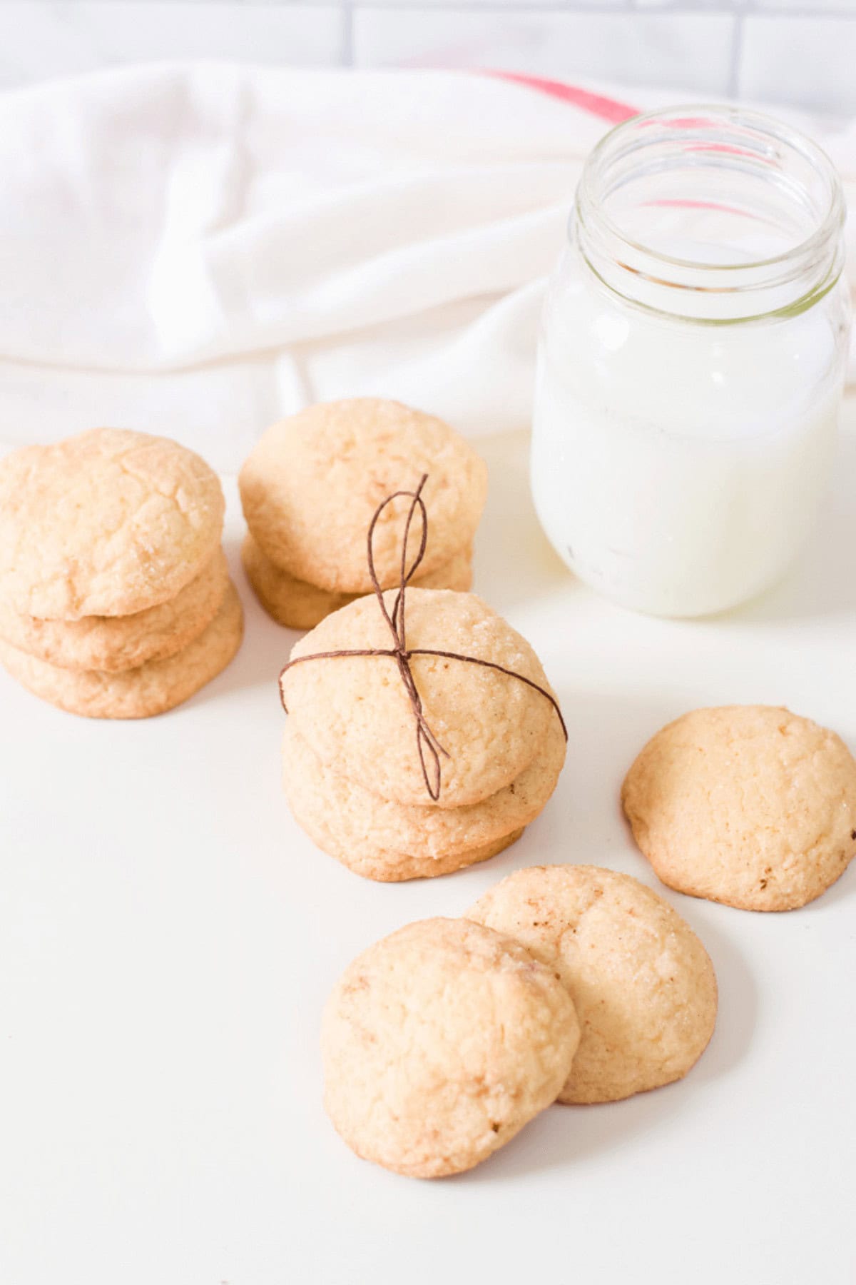 Dessert stacked and tied with twine rest on a white surface beside a glass jar of milk, with additional stacks of cookies nearby.