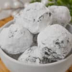 A white bowl filled with Peanut Butter Truffles, delicately dusted in powdered sugar, rests on a wooden tray. White decorative elements and green foliage softly blur in the background.