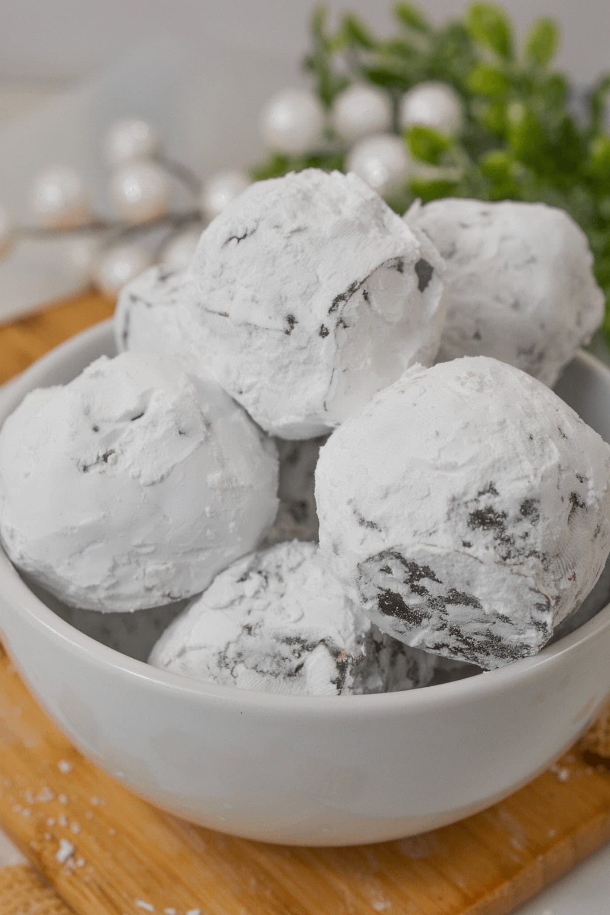 A white bowl filled with Peanut Butter Truffles, delicately dusted in powdered sugar, rests on a wooden tray. White decorative elements and green foliage softly blur in the background.