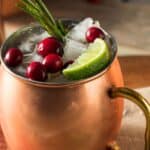 A copper mug filled with ice, cranberries, a lime wedge, a rosemary sprig, and a festive Christmas Moscow Mule sits atop a checked cloth.