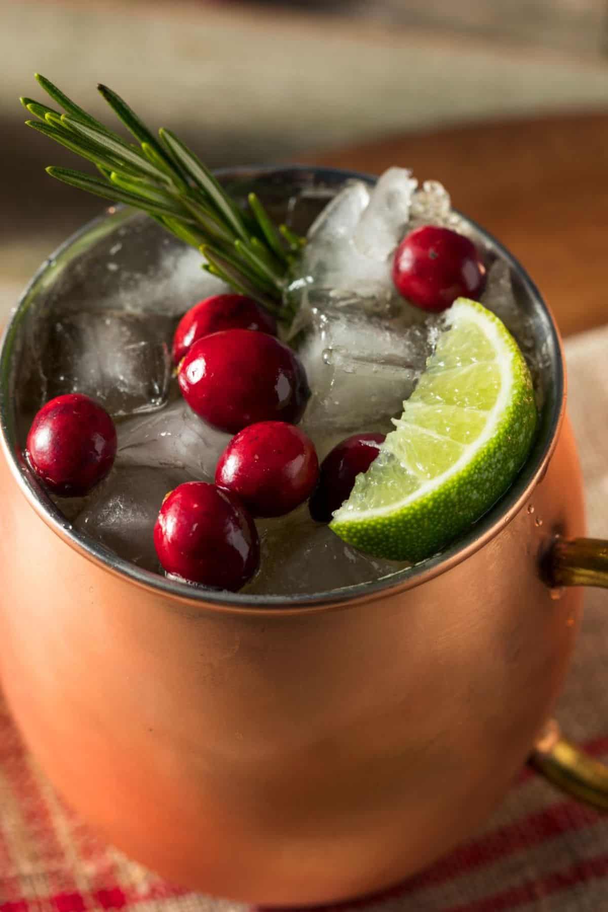 A copper mug filled with ice, cranberries, a lime wedge, and a sprig of rosemary—this Christmas Moscow Mule is served on a red plaid cloth for festive flair.
