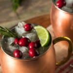 Two copper mugs filled with ice, cranberries, lime slices, and a sprig of rosemary sit on a striped cloth—perfect for serving a festive Christmas Moscow Mule.
