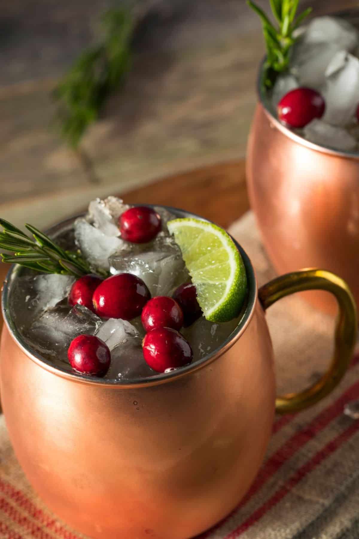 Two copper mugs filled with ice, cranberries, lime slices, and a sprig of rosemary sit on a striped cloth—perfect for serving a festive Christmas Moscow Mule.