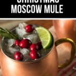 A copper mug filled with a festive Christmas Moscow Mule, garnished with ice, cranberries, a lime slice, and a sprig of rosemary, rests on a striped cloth.
