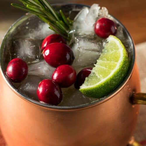 A copper mug filled with ice, cranberries, a lime wedge, and a sprig of rosemary, this festive drink is likely a Christmas Moscow Mule cocktail.