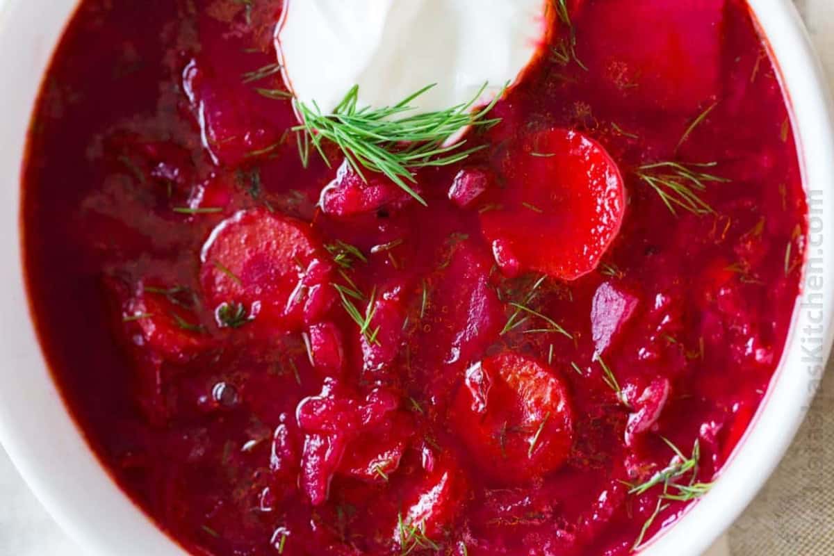 A bowl of red borscht beet soup, a classic Russian food, garnished with fresh dill and a dollop of sour cream. Slices of beets are visible in the reddish broth.