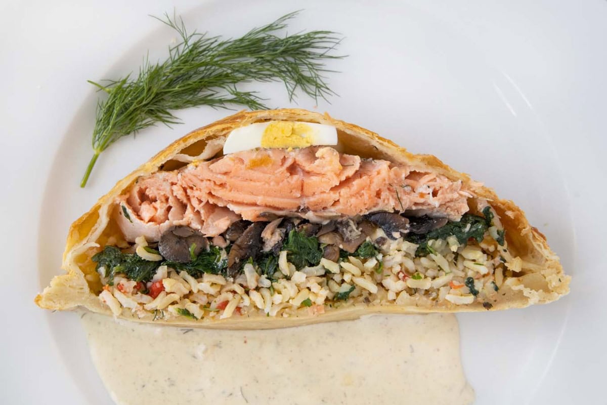 A cross-section of salmon wellington, or coulibiac of salmon, with rice, spinach, and mushrooms inside puff pastry, topped with a slice of boiled egg and dill, served with a creamy sauce—a festive Russian Christmas food classic.