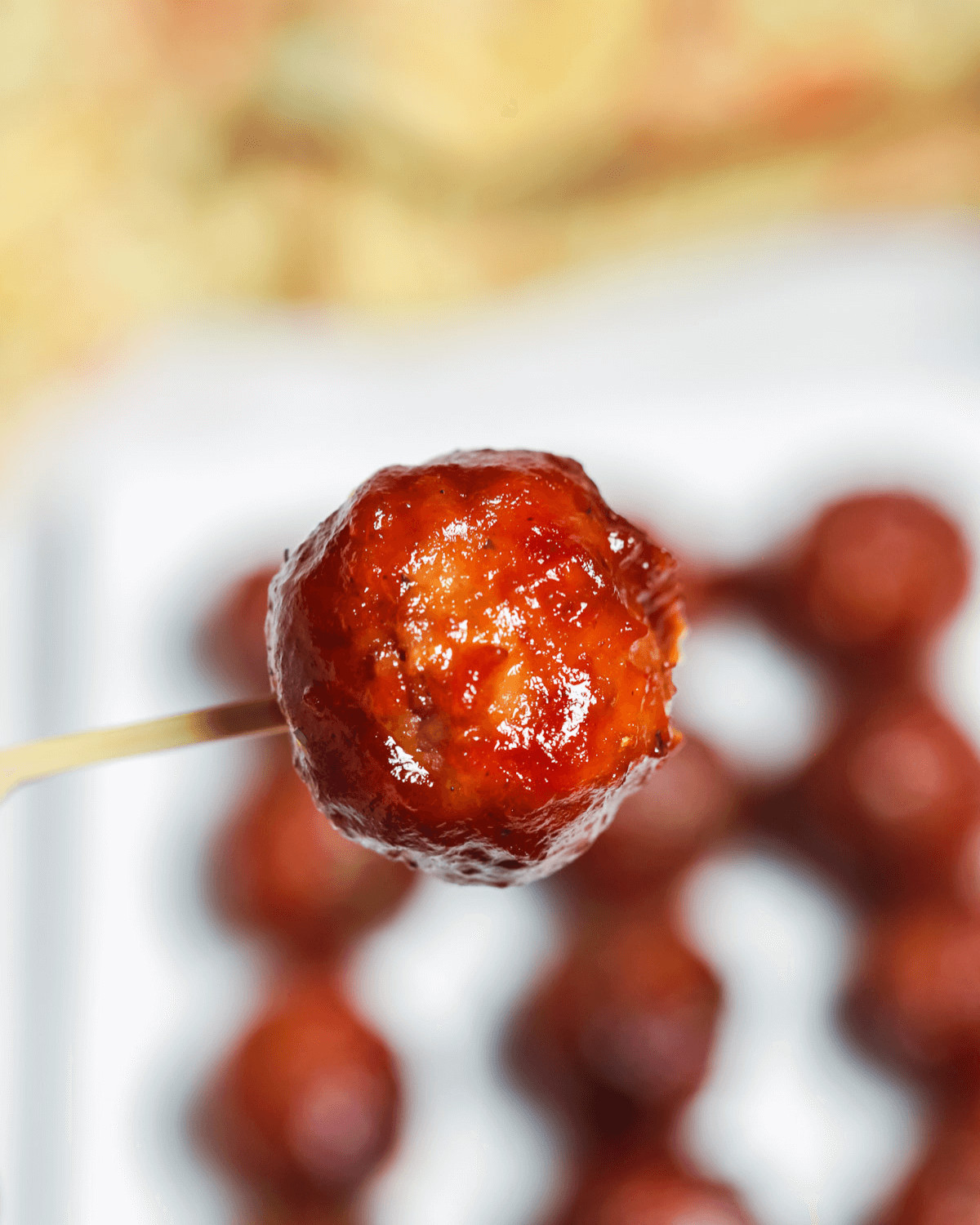 A close-up of a sweet and spicy meatball on a toothpick, with more meatballs in the background.