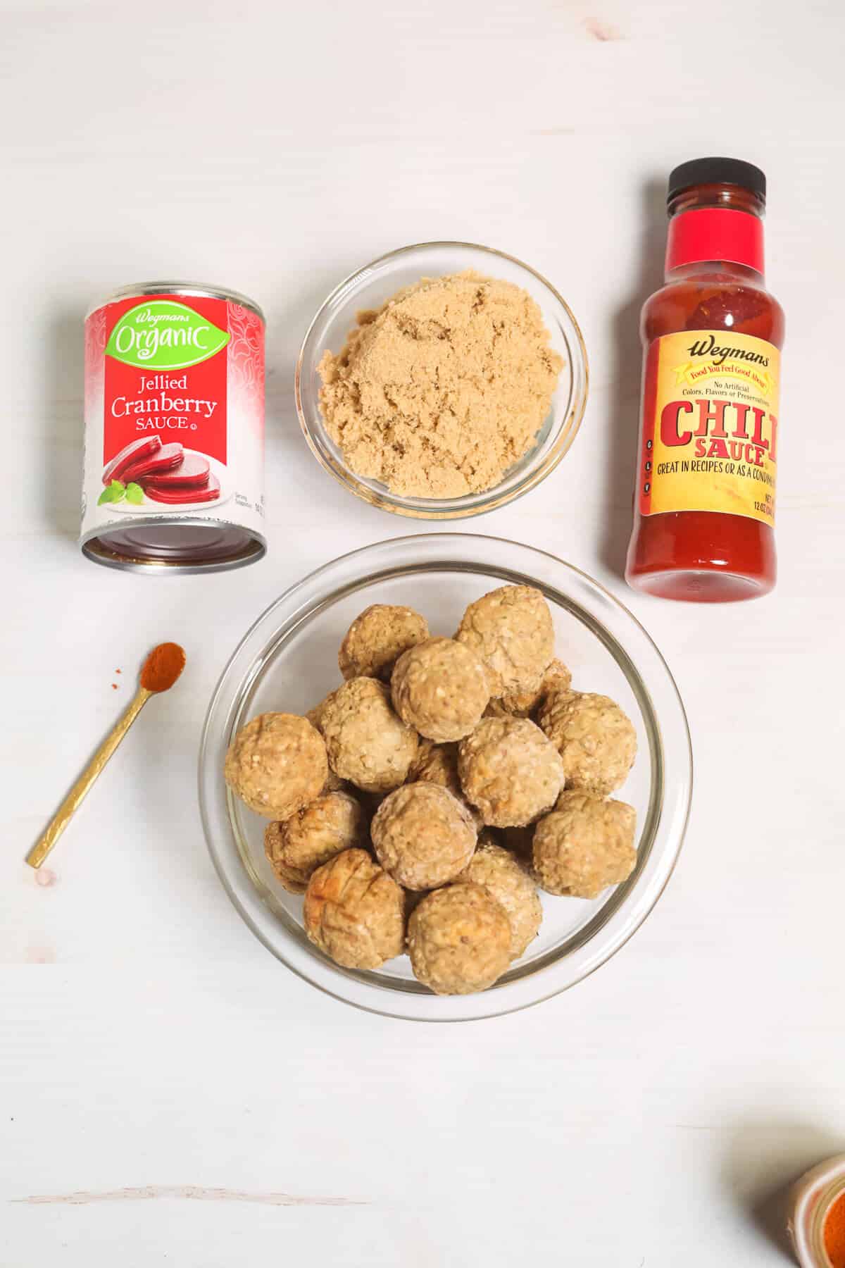 Ingredients for the recipe including canned cranberry sauce, brown sugar, chili sauce, and a bowl of meatballs on a white surface.