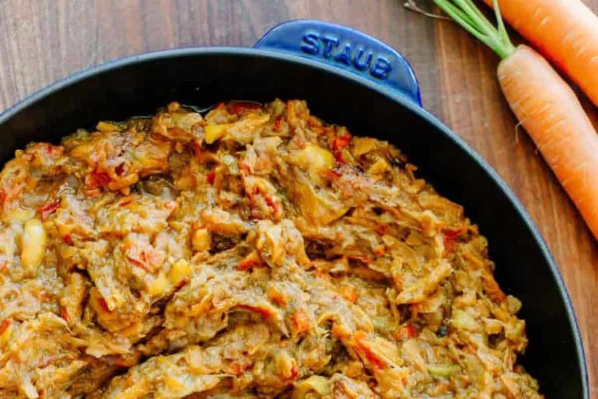 Shredded eggplant and vegetable spread (Baklazhannaia Ikra) in a black Staub dish on a wooden surface, with two carrots and their green tops beside the dish.