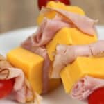 Appetizer skewers with cubes of yellow cheese, slices of ham, and cherry tomatoes artfully arranged on a white plate.