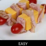 Skewers with cheddar cheese cubes, slices of ham, and cherry tomatoes elegantly displayed on a white plate. Text reads, "Easy Appetizer Skewers.