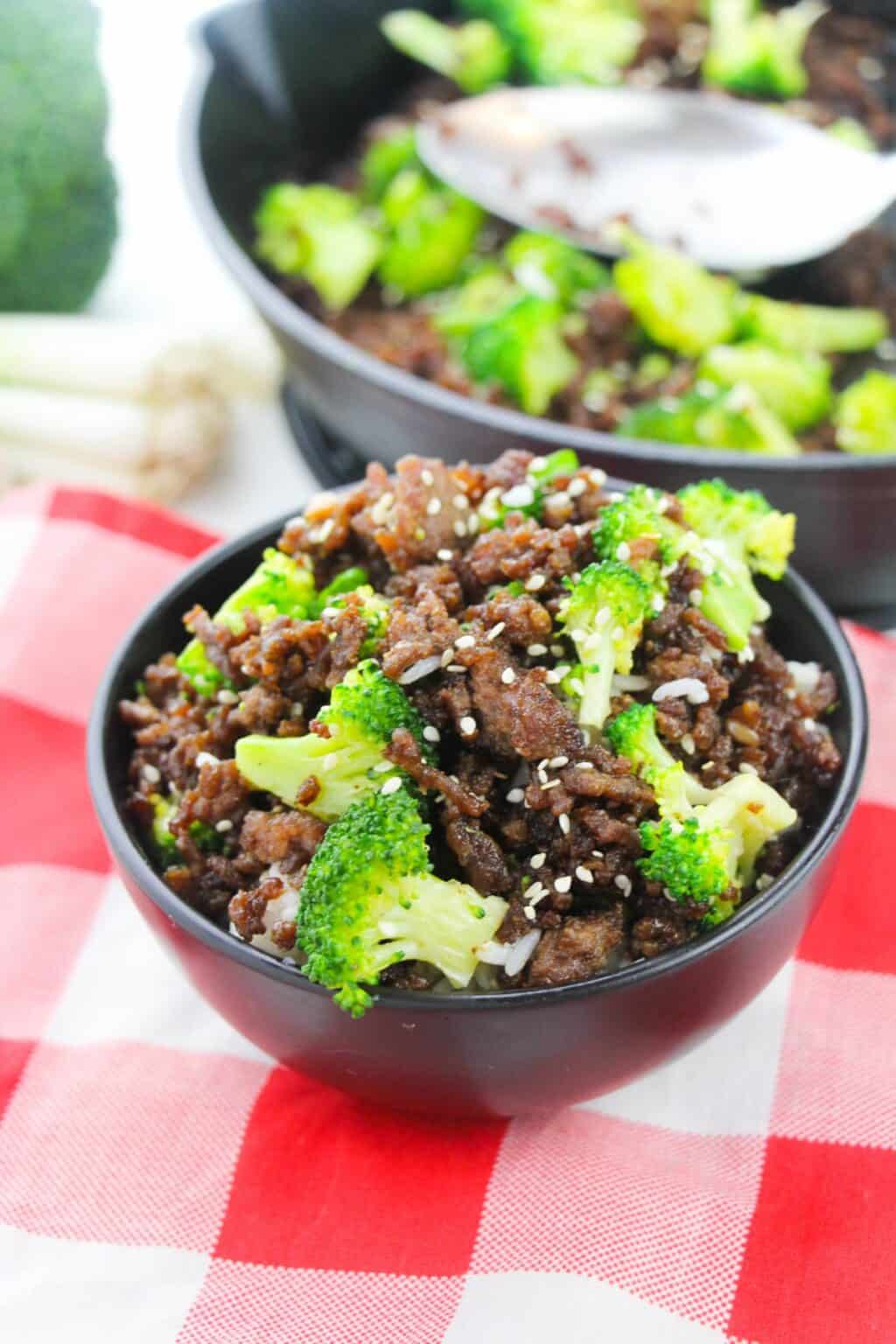 A black bowl filled with Ground Beef and Broccoli, white rice, and sesame seeds sits on a red and white checkered cloth with a similar dish in a pan behind it.