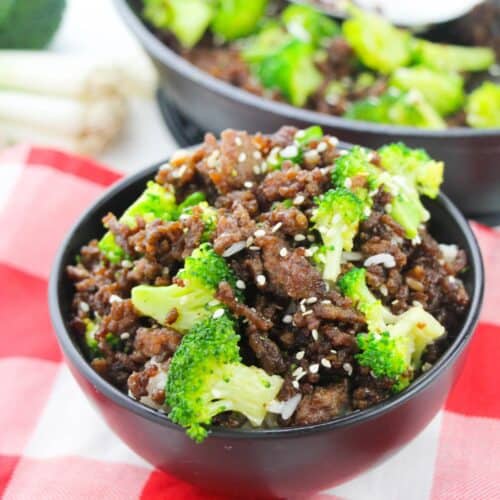 A black bowl filled with Ground Beef and Broccoli, white rice, and sesame seeds sits on a red and white checkered cloth with a similar dish in a pan behind it.