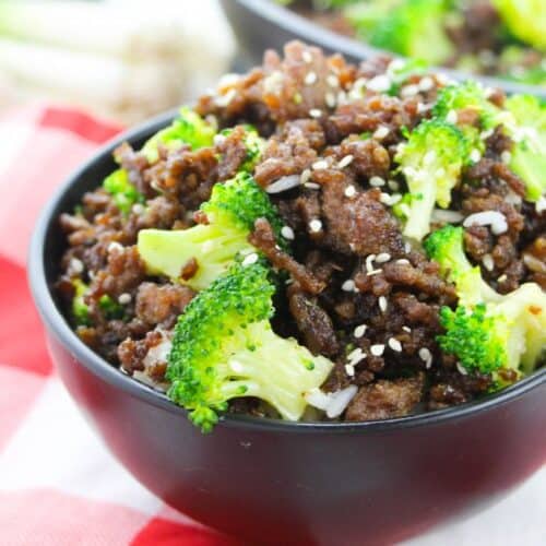 A black bowl filled with flavorful Korean Beef and Broccoli—tender cooked ground beef and fresh broccoli florets, sprinkled with sesame seeds—sits on a red and white checkered cloth.
