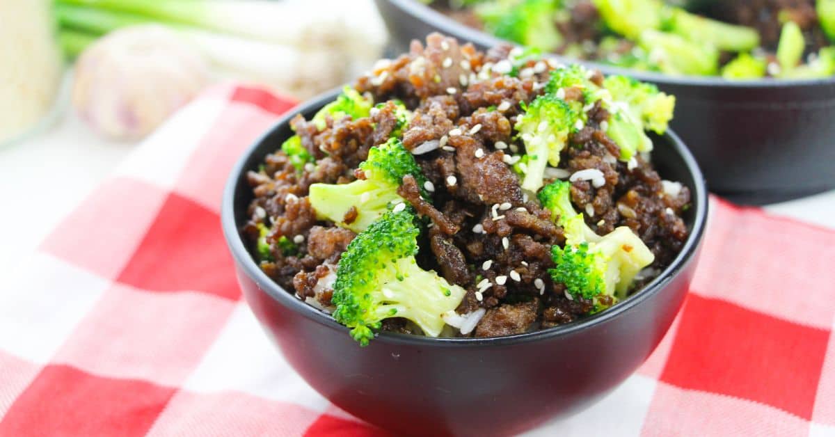 A black bowl filled with flavorful Korean Beef and Broccoli—tender cooked ground beef and fresh broccoli florets, sprinkled with sesame seeds—sits on a red and white checkered cloth.