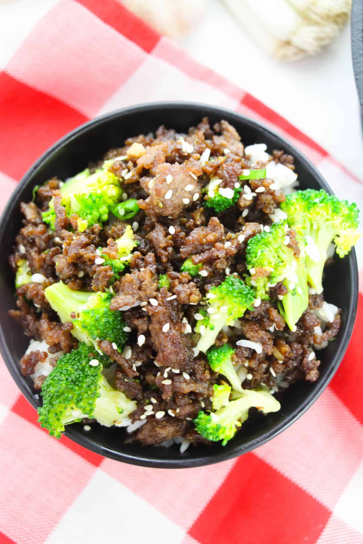 A black bowl filled with Korean Beef and Broccoli, tender broccoli florets, and white rice, topped with sesame seeds, sits on a red and white checkered cloth.