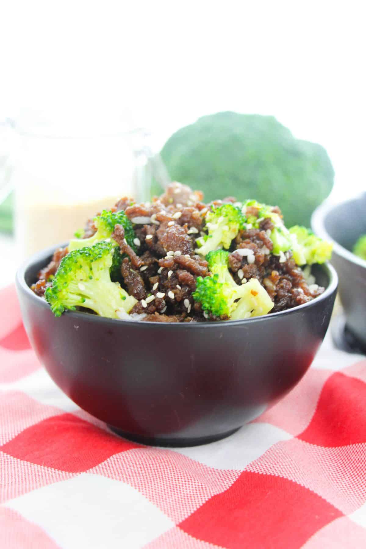 A black bowl filled with Korean Beef and Broccoli, served over rice, sits on a red and white checkered cloth.