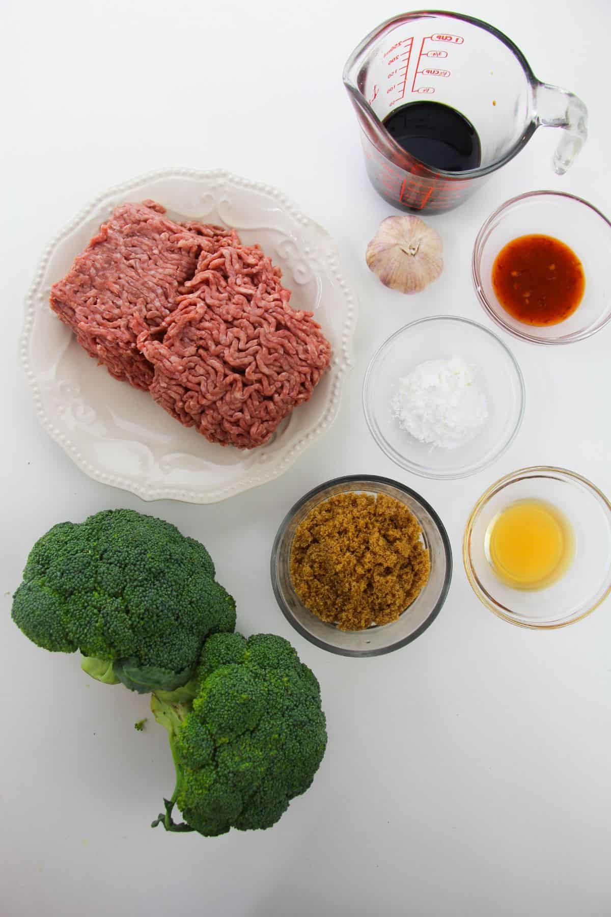 Raw ground beef, broccoli, garlic, brown sugar, soy sauce, a small bowl of red sauce, cornstarch, and a small cup of oil are artfully arranged on a white surface—everything you need to make flavorful Korean Beef and Broccoli at home.