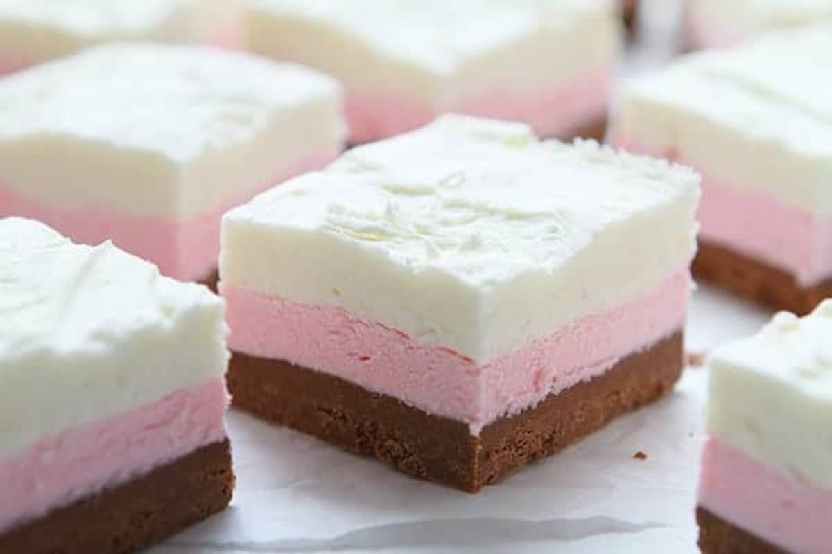 Squares of Neapolitan fudge with three layers—a brown chocolate old fashioned fudge base, a pink raspberry middle layer, and a white chocolate vanilla top—are artfully arranged on a white surface.