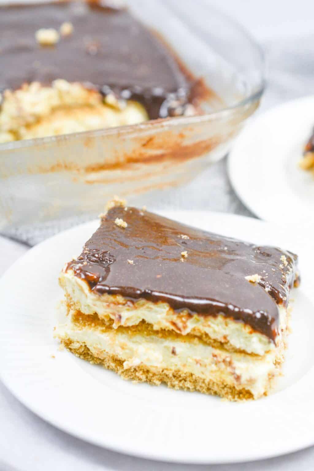 A slice of no bake chocolate eclair cake with a creamy filling and rich chocolate topping sits on a white plate; the rest of the dessert is in a glass dish in the background.