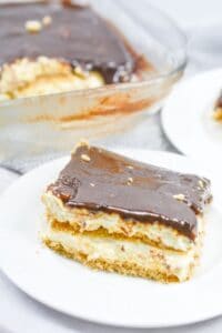 A slice of no bake chocolate eclair cake with a creamy filling and rich chocolate topping sits on a white plate; the rest of the dessert is in a glass dish in the background.