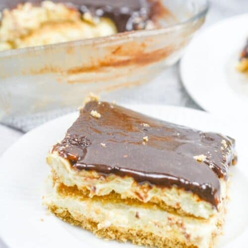 A slice of no bake chocolate eclair cake with a creamy filling and rich chocolate topping sits on a white plate; the rest of the dessert is in a glass dish in the background.