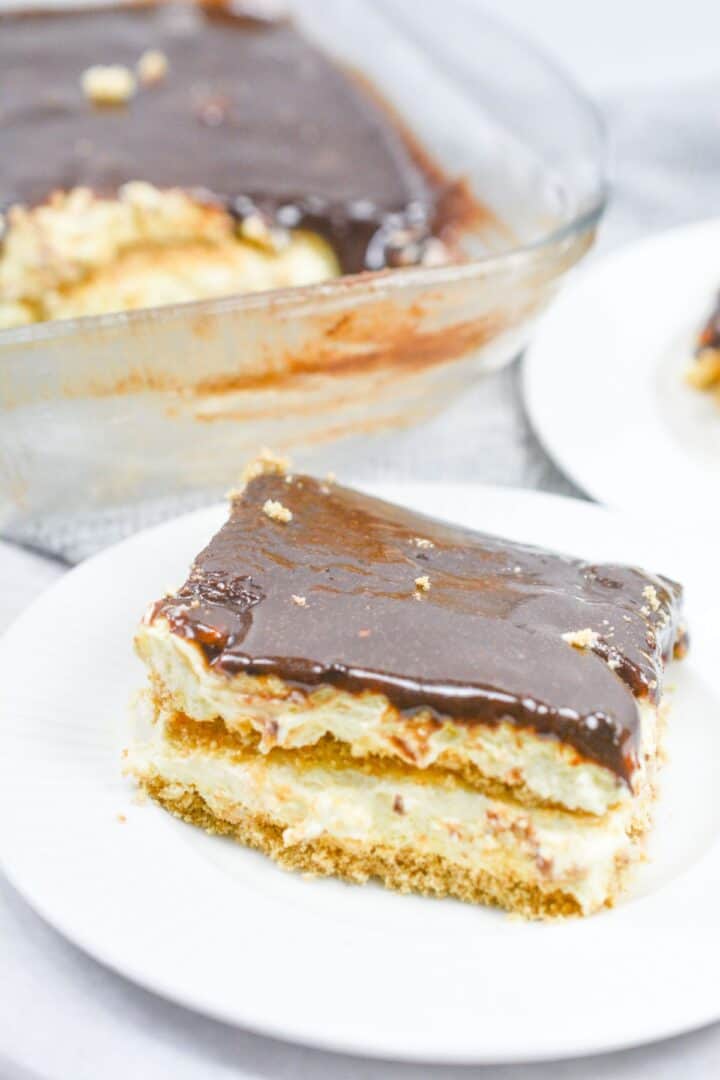 A slice of no bake chocolate eclair cake with a creamy filling and rich chocolate topping sits on a white plate; the rest of the dessert is in a glass dish in the background.
