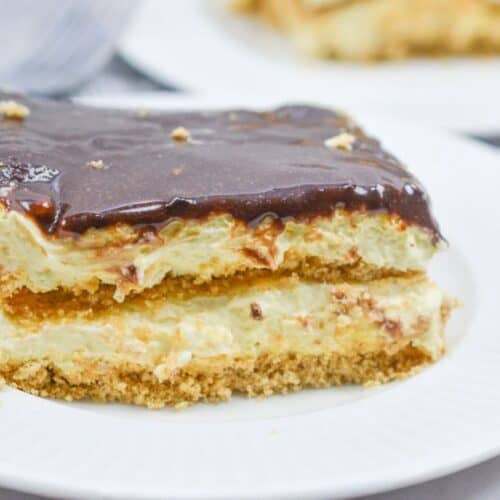 A slice of no bake chocolate eclair cake features a graham cracker crust, creamy filling, and rich chocolate topping on a white plate.