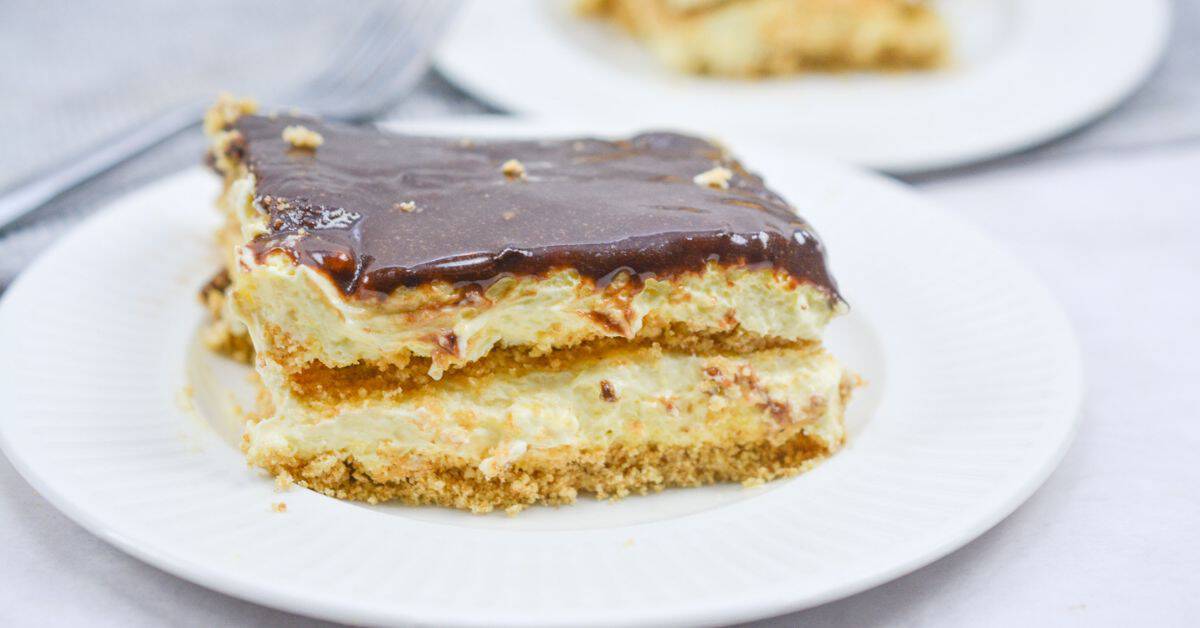 A square slice of no bake chocolate eclair cake with a chocolate topping and creamy filling sits on a white plate, with another slice in the background.