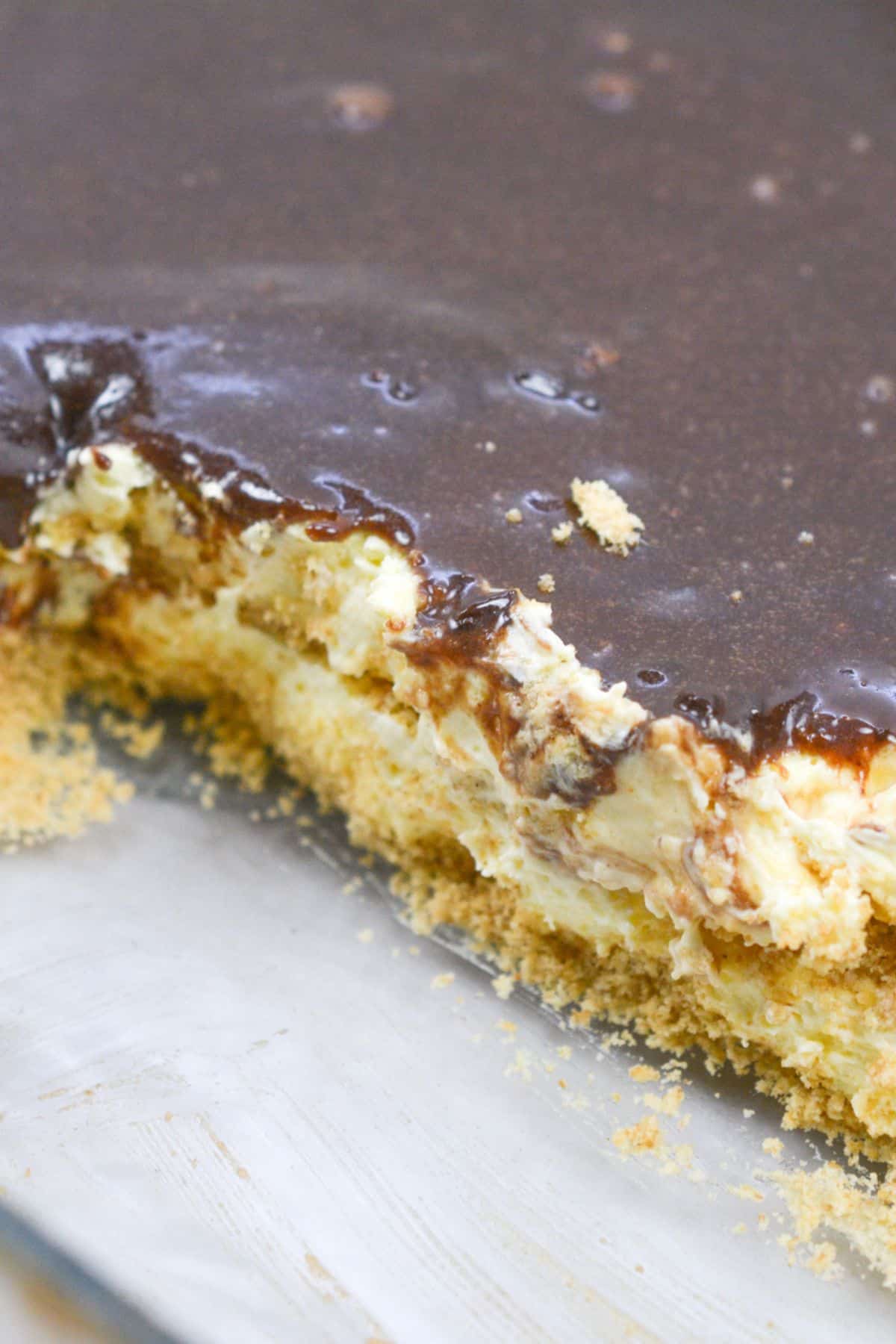 A close-up of a no bake chocolate eclair cake with a graham cracker crust, creamy middle layer, and glossy chocolate topping, with a portion removed to reveal the delicious layers.