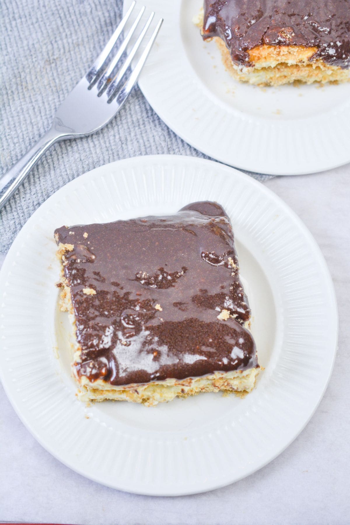 A square piece of no bake chocolate eclair cake with chocolate frosting and cream layers sits on a white plate, next to a fork and another plate holding a similar dessert.