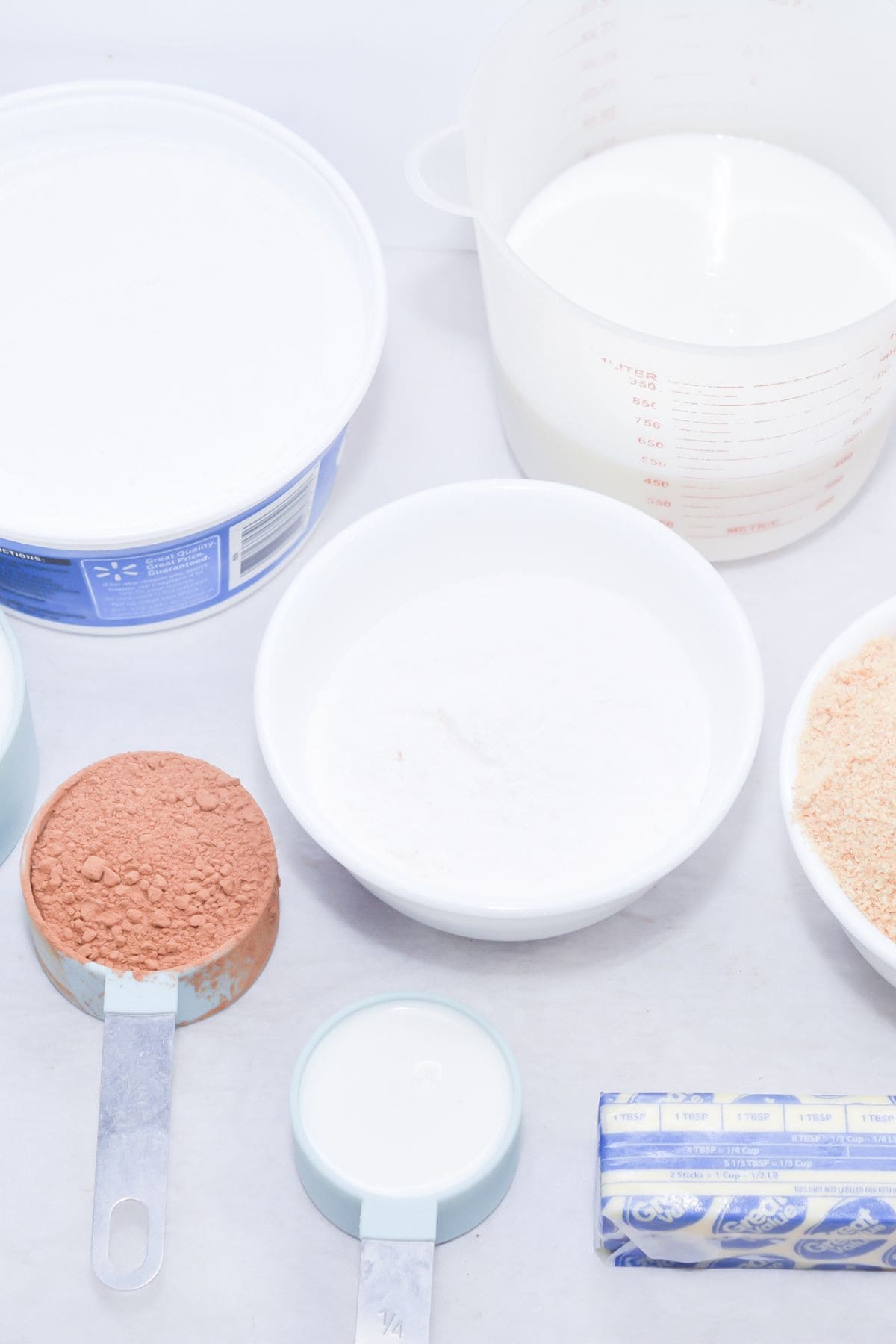 Various baking ingredients for a no bake chocolate eclair cake, including cocoa powder, granulated sugar, butter, milk, and cream, are arranged in bowls, measuring cups, and a jug on a white surface.