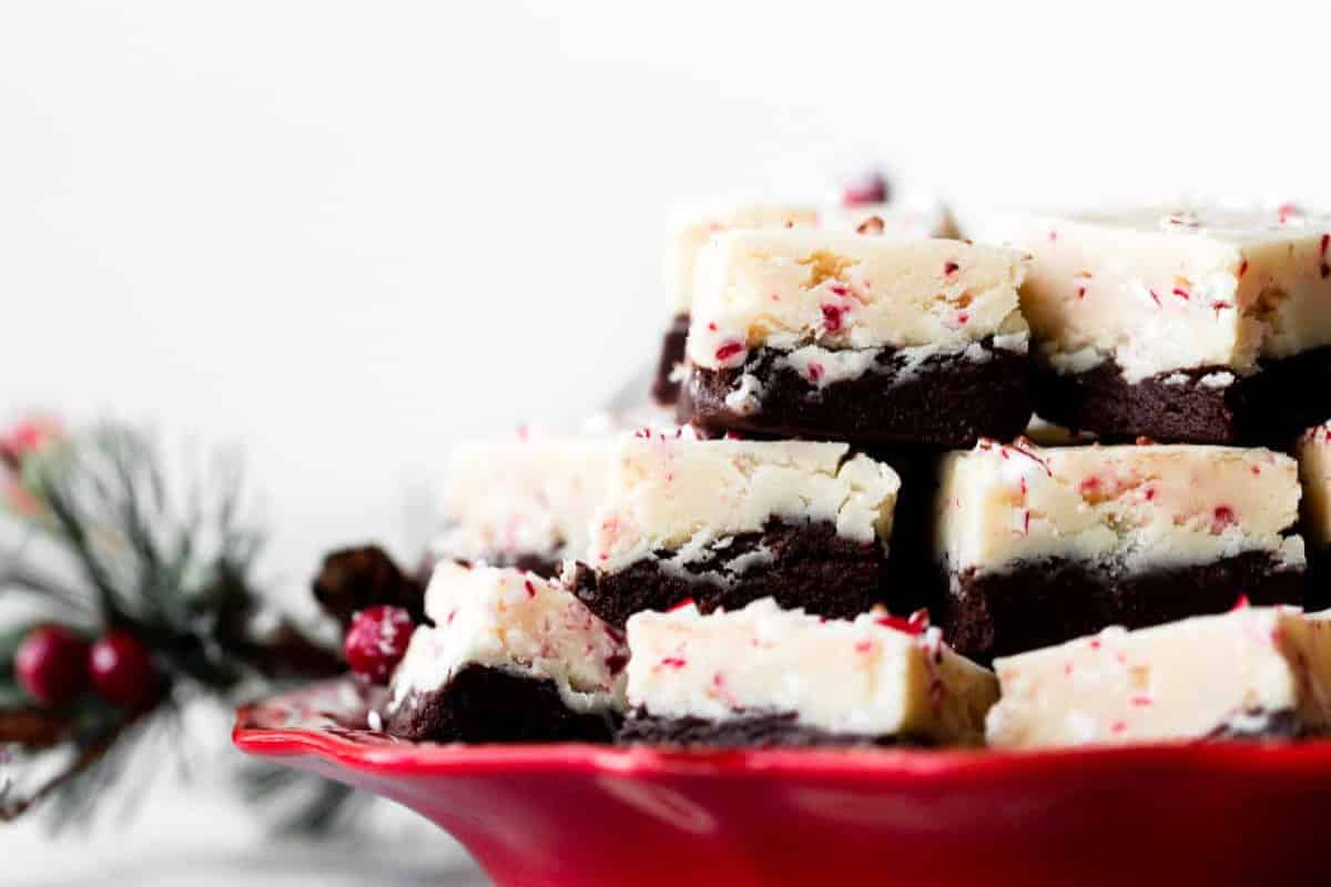 A red plate stacked with layered peppermint bark fudge with holiday decorations in the background.